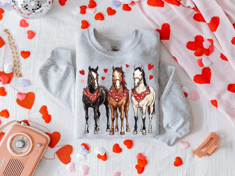 Puede incluir: Una sudadera gris con un gr&aacute;fico de tres caballos con pa&ntilde;uelos. Los caballos son marr&oacute;n, blanco y negro. El fondo es una superficie blanca con corazones rojos.
