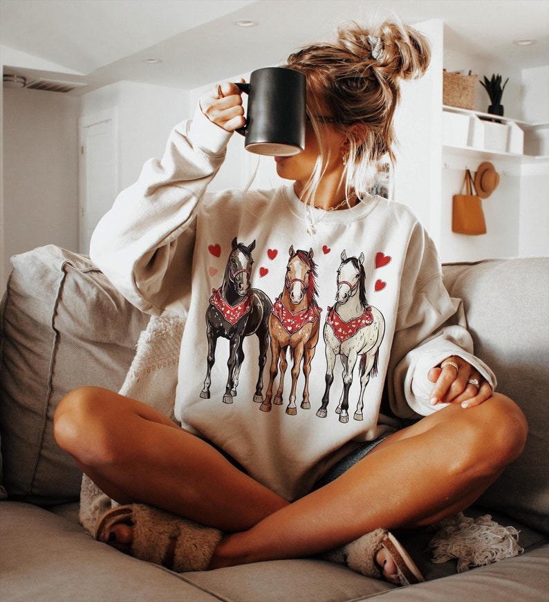 Puede incluir: Una sudadera blanca con un gr&aacute;fico de tres caballos con bandanas rojas y rodeados de corazones rojos. La sudadera la lleva puesta una persona sentada en un sof&aacute;.
