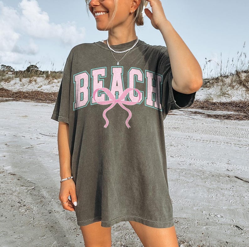 Pu&ograve; includere: Una maglietta oversize verde scuro con un contorno verde chiaro della parola "BEACH" e un fiocco rosa.
