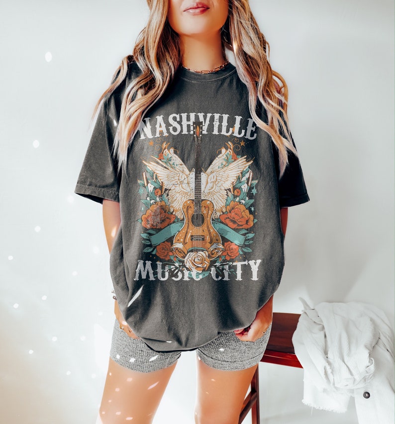 May include: A dark gray t-shirt with a vintage-style graphic of a guitar with wings and flowers. The text "Nashville Music City" is printed on the shirt.