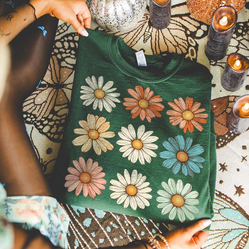 May include: A dark green sweatshirt featuring a colourful daisy print. The daisies are in shades of pink, blue, yellow, and white.