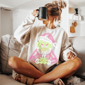 Puede incluir: Sudadera beige con un dise&ntilde;o de ne&oacute;n del Grinch en rosa y verde, con un gorro de Pap&aacute; Noel. Una persona sostiene una taza negra mientras est&aacute; sentada en un sof&aacute; gris. El dise&ntilde;o del Grinch es el punto focal.