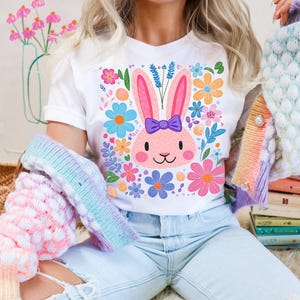 May include: A white t-shirt with a pink bunny rabbit wearing a purple bow and surrounded by colorful flowers. The bunny is the center of the design and is surrounded by a variety of flowers in shades of pink, blue, yellow, and green.