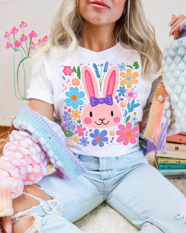 May include: A white t-shirt with a pink bunny rabbit wearing a purple bow and surrounded by colorful flowers. The bunny is the center of the design and is surrounded by a variety of flowers in shades of pink, blue, yellow, and green.