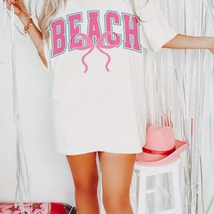 Pu&ograve; includere: Maglietta bianca oversize con una grafica rosa "BEACH" e un fiocco rosa. La maglietta &egrave; indossata da una persona che indossa stivali da cowboy bianchi.