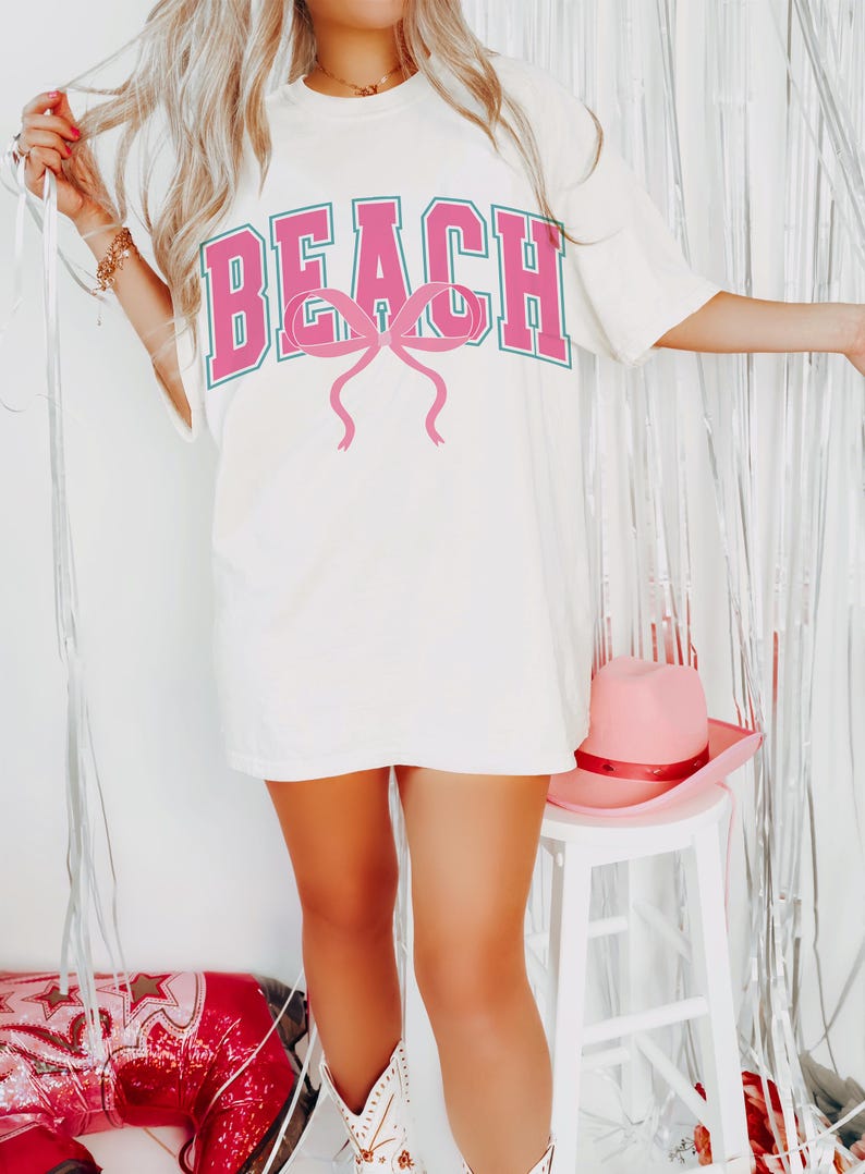 Pu&ograve; includere: Maglietta bianca oversize con una grafica rosa "BEACH" e un fiocco rosa. La maglietta &egrave; indossata da una persona che indossa stivali da cowboy bianchi.