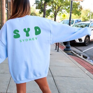 May include: A light blue sweatshirt with a green graphic on the back that says "SYD SYDNEY".