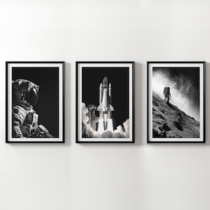 May include: Set of three framed black and white space-themed prints. One print features an astronaut, another a rocket launch, and the third an astronaut climbing a hill. The prints are displayed on a white wall.