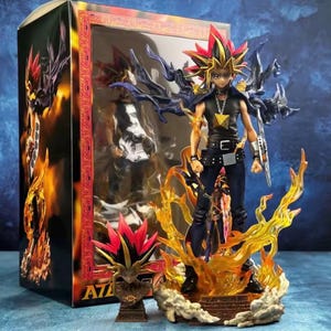 Könnte beinhalten: Yami Yugi Actionfigur aus der Anime-Serie Yu-Gi-Oh!. Die Figur posiert auf einem feurigen Sockel mit schwarzer und goldener Kleidung, stacheligen Haaren und einem Millenniums-Puzzle-Anhänger. Die Figur wird in einer Box mit Fenster präsentiert.