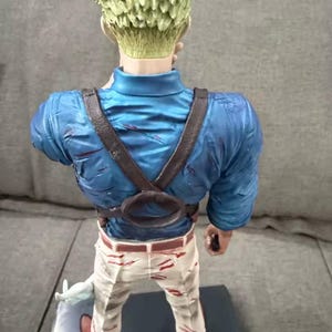May include: A figurine of a character with light green hair, wearing a blue shirt, brown suspenders, and white pants with red accents. The figure is holding a white object and is standing on a dark base.
