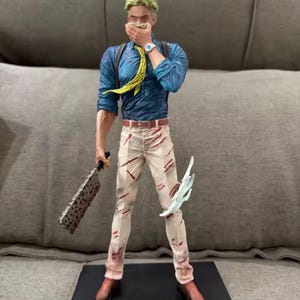 May include: A detailed figurine of a character in a blue shirt, yellow tie, and white pants with red accents. The figure holds a cleaver and a translucent blue energy effect. The figure is standing on a black base.
