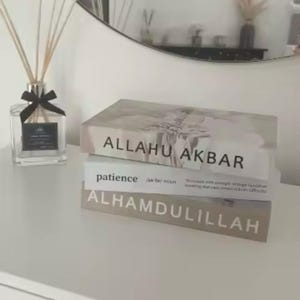 Puede incluir: Una pila de libros decorativos con las frases "ALLAHU AKBAR", "patience" y "ALHAMDULILLAH" impresas en los lomos. A la izquierda, un frasco difusor de vidrio transparente con cañas y una cinta negra.