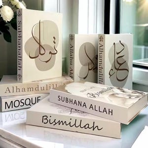Può includere: Una collezione di libri decorativi in varie dimensioni e orientamenti. I libri sono bianco sporco con calligrafia marrone e forme astratte. Il testo include "Subhanallah", "Bismillah" e "Alhamdulillah". I libri sono disposti su una superficie riflettente.