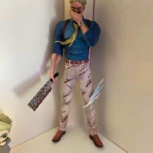 May include: A detailed figurine of a character in a blue shirt, tan pants with red markings, and brown shoes. The figure is holding a large, serrated weapon in one hand and a blue, stylized energy effect in the other. A yellow and black patterned tie is visible.