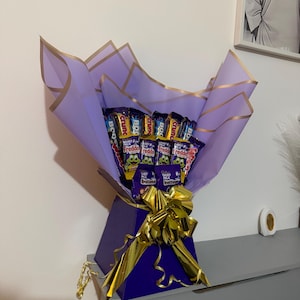 May include: A chocolate bouquet presented in a purple gift box with a gold bow. The arrangement features various chocolate bars, wrapped in purple and gold paper. A sweet gift idea.