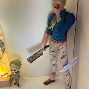 May include: A detailed figurine of a character from an anime series, wearing a blue shirt, suspenders, and white pants with red streaks. The character holds a large cleaver and has a yellow patterned tie. A smaller figurine is also present.