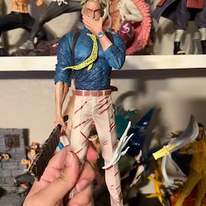 May include: A detailed figurine of a character in a blue shirt, tan pants with red streaks, and a yellow patterned tie. The figure is holding a weapon and has a watch on its wrist. Other figurines are visible on a shelf in the background.