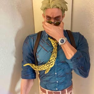 May include: A figurine of a character wearing a blue shirt, brown suspenders, and a yellow patterned tie. The character is wearing sunglasses and a silver watch. The figurine is a collectible item.