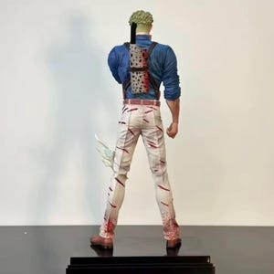 May include: A figurine of a character from an anime series, viewed from the back. The figure wears a blue shirt, white pants with red streaks, and brown boots. A sheath with swords is strapped to the back. The figure has green hair.