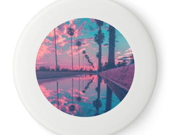 May include: White frisbee featuring a circular image of a sunset over a road with palm trees. The sky is pink, purple, and blue, reflected in the water. A fun outdoor toy for playing in the park.