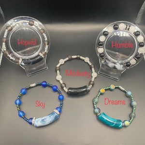 Könnte beinhalten: Vier Armbänder mit unterschiedlich farbigen Perlen und Charms. Die Armbänder sind mit "Hopeful", "Humble", "Modesty" und "Dreams" beschriftet.