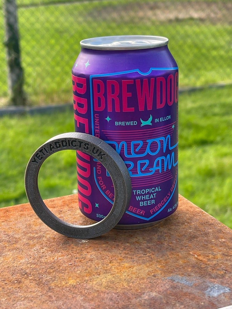 May include: A purple and pink can of Brewdog Neon Dream Tropical Wheat Beer with a black rubber ring around it. The can has the text "Brewdog" in pink and "Neon Dream" in blue. The can also has the text "Tropical Wheat Beer" and "Fiercely Defiant" in blue. The can is sitting on a rusty surface.