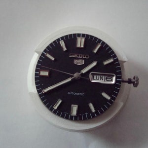 Puede incluir: Una esfera de reloj negra con marcas blancas y un bisel plateado. La esfera del reloj tiene las palabras "SEIKO" y "AUTOMATIC" impresas. La ventana de fecha muestra "LUN 6".