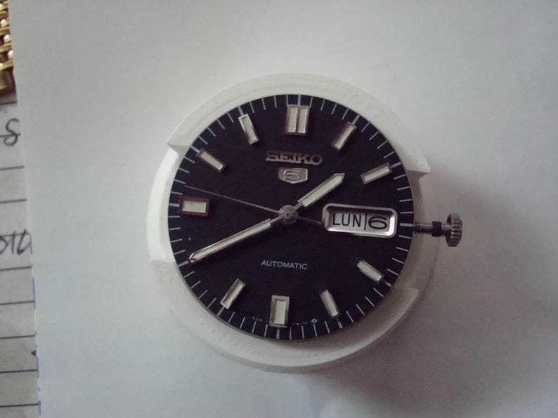 Puede incluir: Una esfera de reloj negra con marcas blancas y un bisel plateado. La esfera del reloj tiene las palabras "SEIKO" y "AUTOMATIC" impresas. La ventana de fecha muestra "LUN 6".