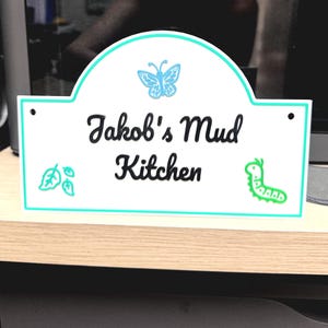 May include: White sign with a teal border and the text "Jakob's Mud Kitchen" in black script. A teal butterfly and a green caterpillar are above and to the right of the text. A teal leaf design is on the left.