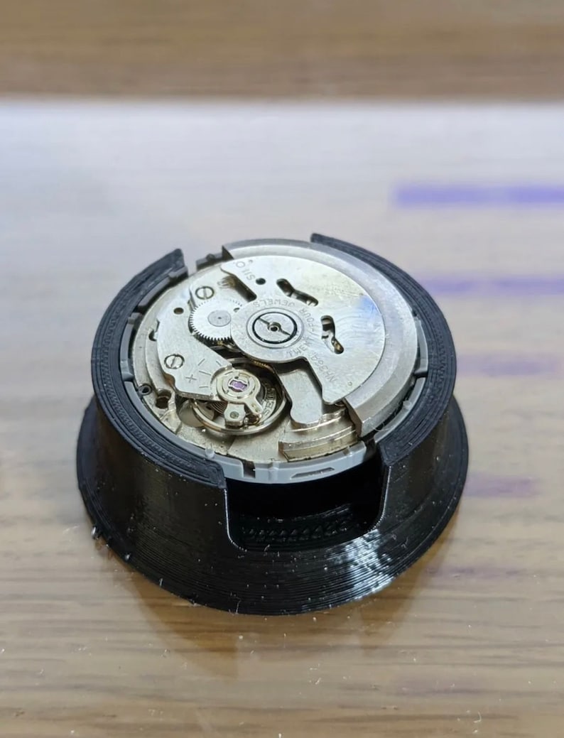 Puede incluir: Un movimiento de reloj plateado con engranajes intrincados y una base de pl&aacute;stico negro.