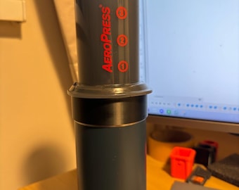 Könnte beinhalten: Eine graue AeroPress Kaffeemaschine mit einer schwarzen und blauen Yeti-Reiseflasche. Die AeroPress hat rote Zahlen 1, 2 und 3 an der Seite.