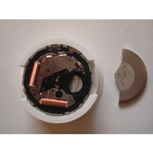 Puede incluir: Primer plano de un movimiento de reloj con bobinas de color cobre y el texto "SM 230" visible. El movimiento est&aacute; en una caja de pl&aacute;stico blanco con un anillo gris alrededor del borde. Tambi&eacute;n se ve una pieza de metal plateada separada.