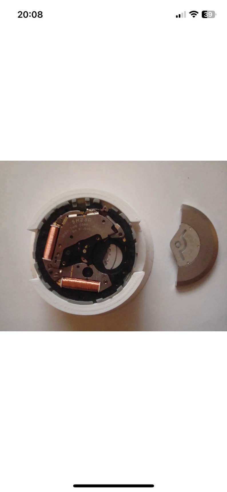 Puede incluir: Primer plano de un movimiento de reloj con bobinas de color cobre y el texto "SM 230" visible. El movimiento est&aacute; en una caja de pl&aacute;stico blanco con un anillo gris alrededor del borde. Tambi&eacute;n se ve una pieza de metal plateada separada.