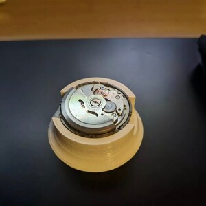 Puede incluir: Primer plano de un movimiento de reloj en un soporte de pl&aacute;stico blanco. El movimiento es plateado con marcas negras y un rotor de color cobre.
