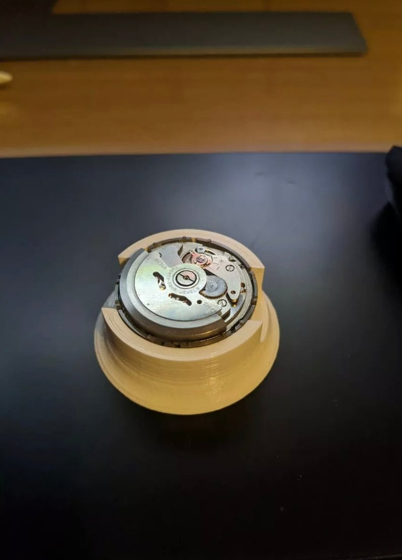 Puede incluir: Primer plano de un movimiento de reloj en un soporte de pl&aacute;stico blanco. El movimiento es plateado con marcas negras y un rotor de color cobre.