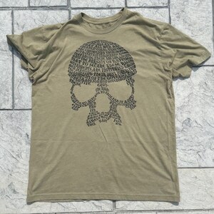 Puede incluir: Una camiseta verde con un gráfico de calavera negra. La calavera está formada por palabras, incluyendo "vida, libertad y la búsqueda de la felicidad".