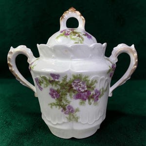 Puede incluir: Azucarero de porcelana blanca con tapa, con un diseño floral de flores moradas y hojas verdes. Las asas y los detalles de la tapa están adornados con ribetes dorados. El cuenco tiene un borde festoneado y una base decorativa.