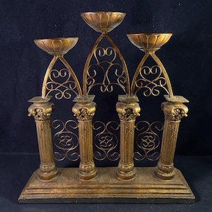 Op de afbeelding: Een goudkleurige, sierlijke kandelaar met vijf kaarsbekers. De basis heeft vijf decoratieve zuilen met ingewikkelde details. De kandelaar heeft een decoratief, gebogen filigraanontwerp op de achtergrond.