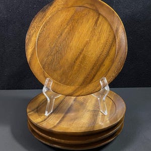 May include: A set of four wooden plates with a natural wood grain finish. The plates are round with a slightly raised edge. One plate is displayed on a clear plastic stand, with the other three stacked below. The plates are a warm brown colour.