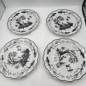 Può includere: Un set di quattro piatti rotondi in ceramica bianca con bordi smerlati, ognuno caratterizzato da una stampa nera di un paesaggio con alberi, un nido e uccelli in volo. Il disegno è centrato sul piatto con un bordo decorativo di rami.