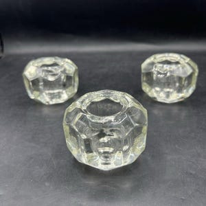 May include: Three clear glass candle holders with a faceted, multi-sided geometric design. Each piece features a circular opening at the top and a thick, transparent base, arranged in a triangular formation on a dark surface.
