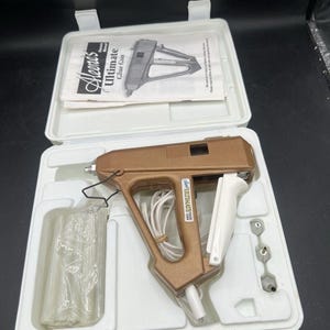 Op de afbeelding: Een bronskleurig Aleene's Ultimate lijmpistool met een witte trekker en stroomkabel, in een witte kunststof koffer. De set bevat een handleiding, drie metalen mondstukken en een doorzichtige kunststof huls voor lijmsticks.