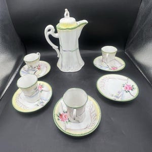 May include: A white porcelain tea set featuring a tall teapot with a lid and four matching cups and saucers. Each piece is decorated with hand-painted pink roses, green leaf accents, and a thin green dotted border along the edges of the items.