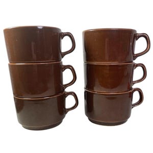 Könnte beinhalten: Sechs gestapelte braune Keramik-Kaffeetassen mit gebogenen Griffen. Die Tassen haben eine einfarbige, dunkelbraune Farbe und scheinen aus Steinzeug zu sein. Die Tassen sind in zwei separaten Stapeln zu je drei Stück gestapelt.