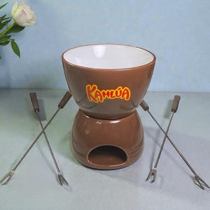 Può includere: Un set per fonduta in ceramica marrone con la scritta "Kahlua" in lettere arancioni e gialle. Il set include una pentola con interno bianco e quattro forchette da fonduta in metallo con manici in legno. Ideale per una serata.