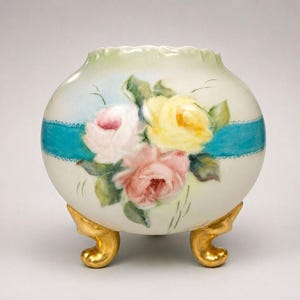 May include: A round porcelain vase with a scalloped rim, featuring a hand-painted bouquet of pink and yellow roses. A horizontal turquoise blue ribbon band encircles the center. The vase is supported by three ornate, metallic gold feet.
