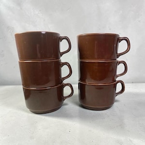 May include: Six brown ceramic coffee cups with handles, stacked in two groups of three. The cups are a solid, warm brown colour and have a simple, classic design. The background is a neutral grey.
