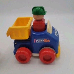 May include: A blue and yellow toy dump truck with red wheels. The truck has a yellow bed, and a red and tan figure wearing a green hat is in the cab. The text "Push'n Go" is on the side.