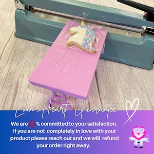 Puede incluir: Una mesa rosa con una galleta de unicornio encima. La mesa se est&aacute; sellando en una bolsa de pl&aacute;stico con una selladora t&eacute;rmica. El texto "Lion Heart Guarantee" es visible en la imagen. El texto "We are 100% committed to your satisfaction. If you are not completely in love with your product please reach out and we will refund your order right away." tambi&eacute;n es visible en la imagen.