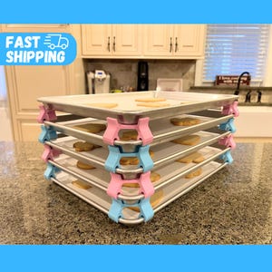 May include: Four silver baking sheets are stacked on top of each other. The sheets are held together by pink and blue plastic clips. The sheets are on a light brown countertop.